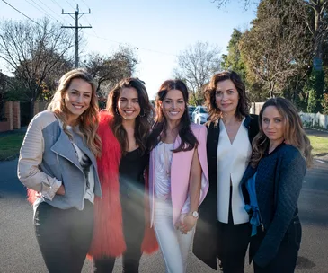 Five women smiling and standing together on a suburban street with trees and houses in the background.