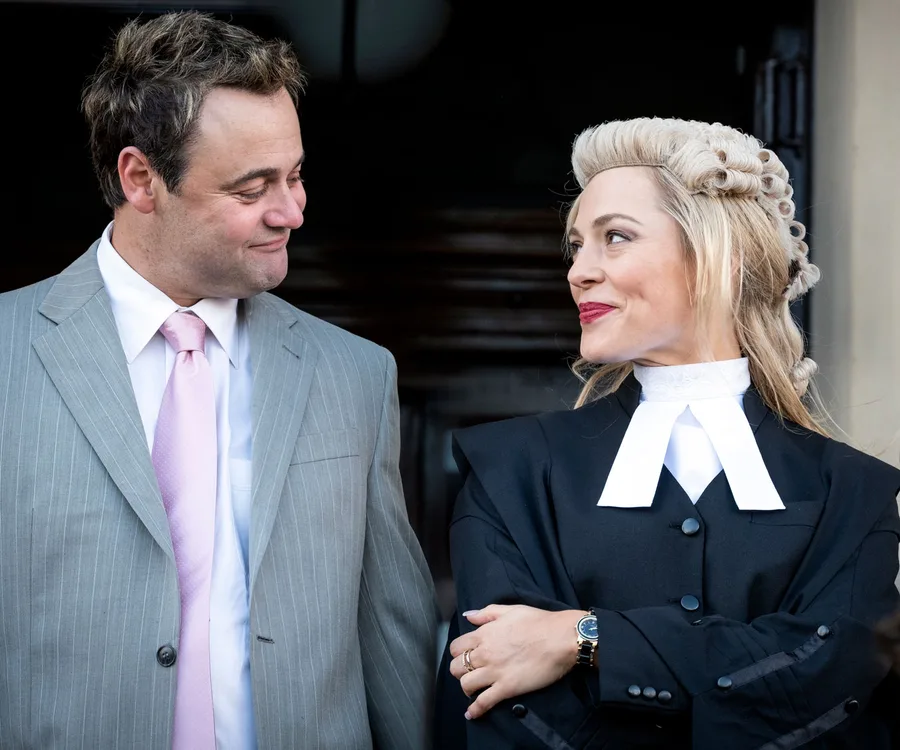 Two actors dressed as a barrister and a man in a suit share a knowing glance, both smiling warmly.