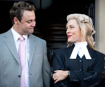 Two actors dressed as a barrister and a man in a suit share a knowing glance, both smiling warmly.