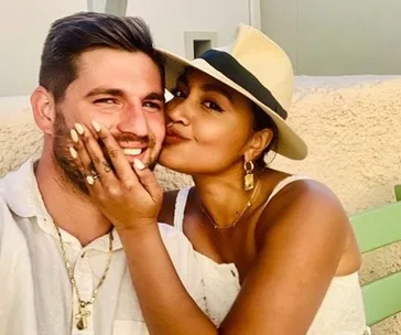 Couple smiling and posing, woman showing engagement ring while kissing man's cheek.