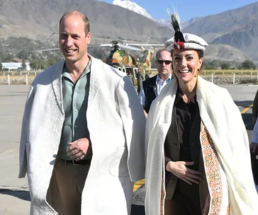 A couple wearing traditional Pakistani attire, including robes and a feathered hat, standing outside with a helicopter in the background.
