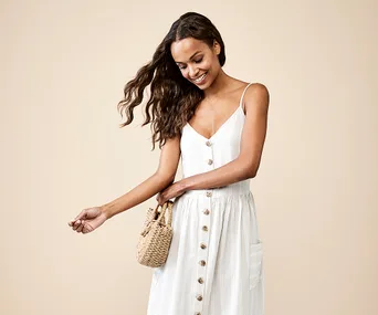 Woman in a white buttoned summer dress holding a straw handbag, smiling against a beige background.