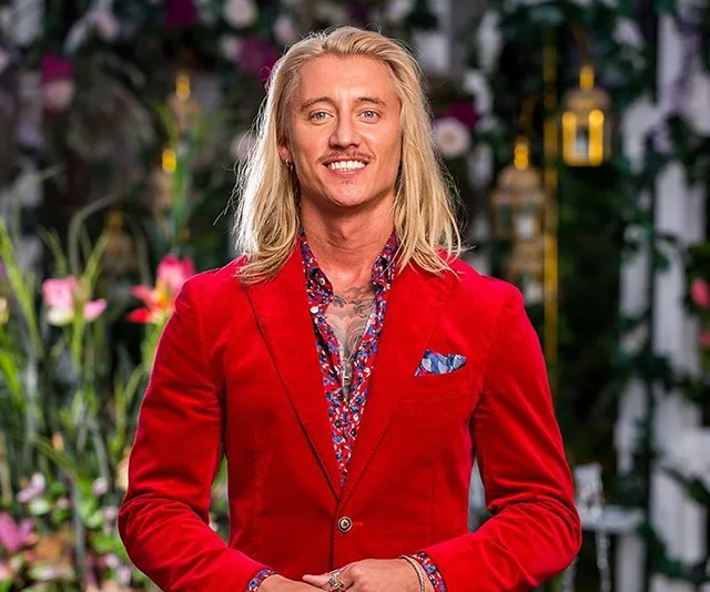 Man in a red suit with a floral shirt stands in a garden setting, smiling at the camera.