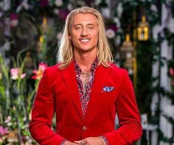 Man in a red suit with a floral shirt stands in a garden setting, smiling at the camera.