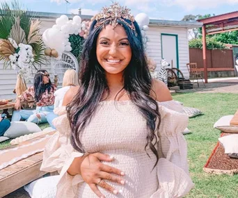 Pregnant woman smiling, wearing a crown and off-shoulder dress, holding her belly at an outdoor event with balloons.