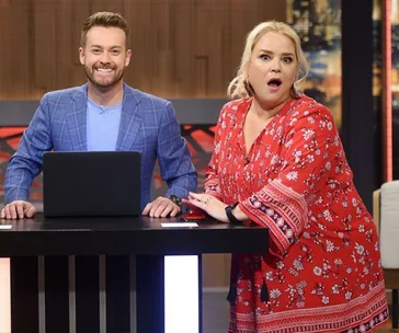 Two radio show hosts, one smiling at a laptop, the other looking surprised, on a studio set.