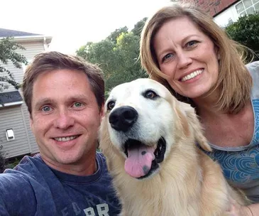 A smiling couple takes a selfie with their happy golden retriever outside.