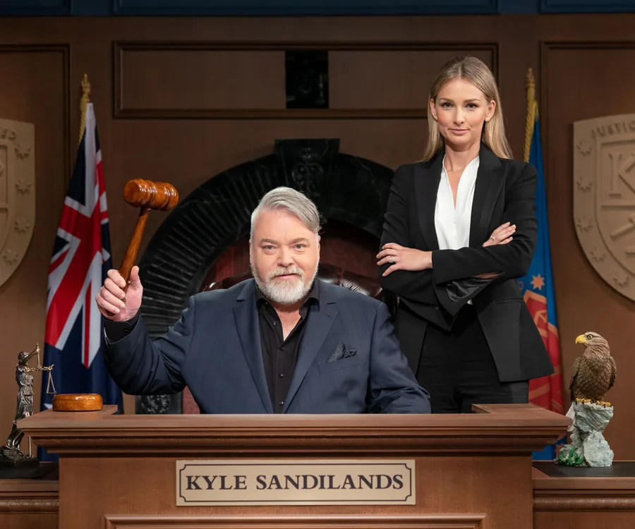 Kyle Sandilands as a judge with a gavel, Anna Heinrich stands beside him in courtroom setting.