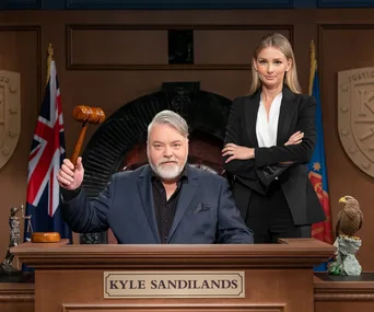 Kyle Sandilands as a judge with a gavel, Anna Heinrich stands beside him in courtroom setting.