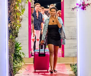 Woman leaving "Love Island" villa with suitcase, smiling, while others look on from doorway.