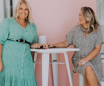 Two women in stylish plus-size dresses enjoying a casual moment by a pink wall.