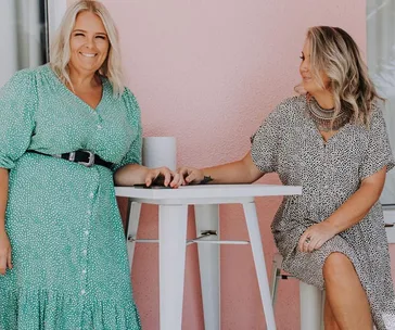 Two women in stylish plus-size dresses enjoying a casual moment by a pink wall.