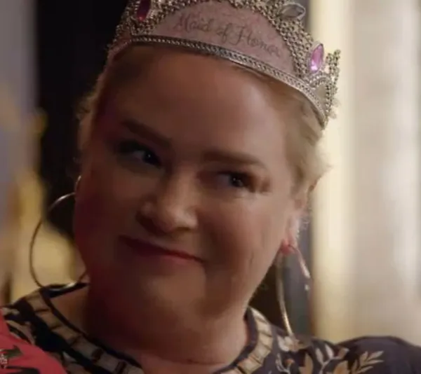 Woman wearing a tiara that says "Maid of Honor" with large hoop earrings, smiling subtly in a warmly lit room.