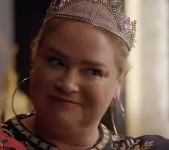 Woman wearing a tiara that says "Maid of Honor" with large hoop earrings, smiling subtly in a warmly lit room.