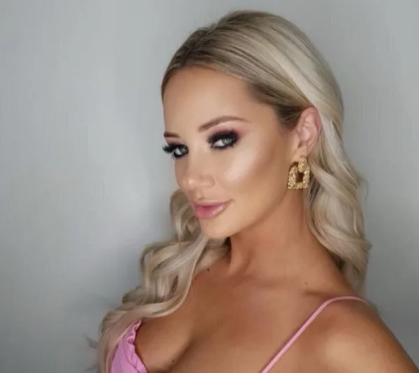 Blonde woman with curled hair and elegant earrings, wearing pink makeup and a pink top, looking over her shoulder.