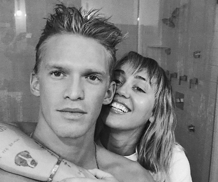A grayscale image of two people smiling closely together, one with short hair and the other with longer, wavy hair.