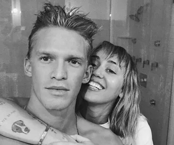 A grayscale image of two people smiling closely together, one with short hair and the other with longer, wavy hair.