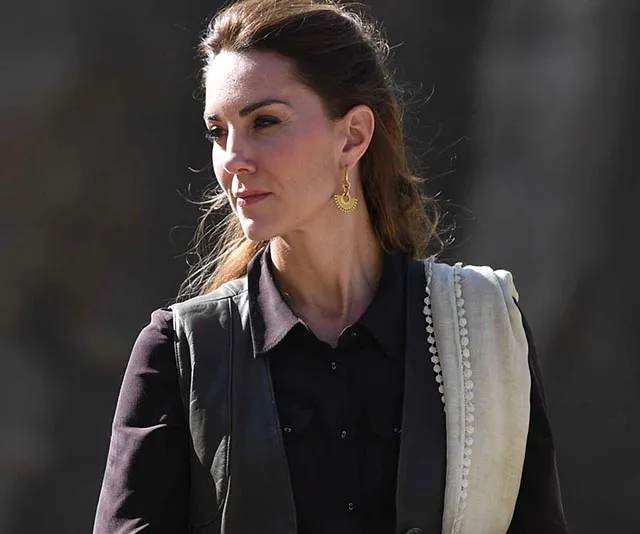 A woman in a black shirt dress with a vest, wearing gold earrings, looks to the side outdoors.