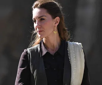 A woman in a black shirt dress with a vest, wearing gold earrings, looks to the side outdoors.