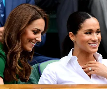 Women seated, smiling at an event, with one wearing a green outfit and the other in white, both with styled hair.