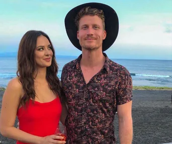 A couple standing on a beach, with the woman in a red dress and the man in a patterned shirt and hat, smiling at the camera.