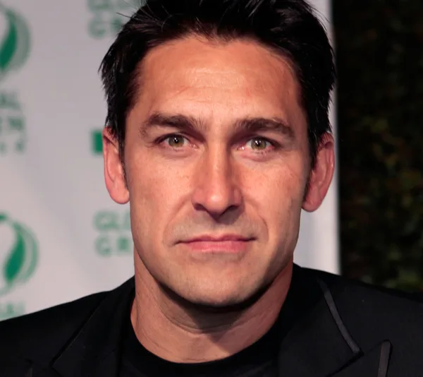 A man in a dark suit stands against a backdrop with green text and logos, looking directly at the camera.