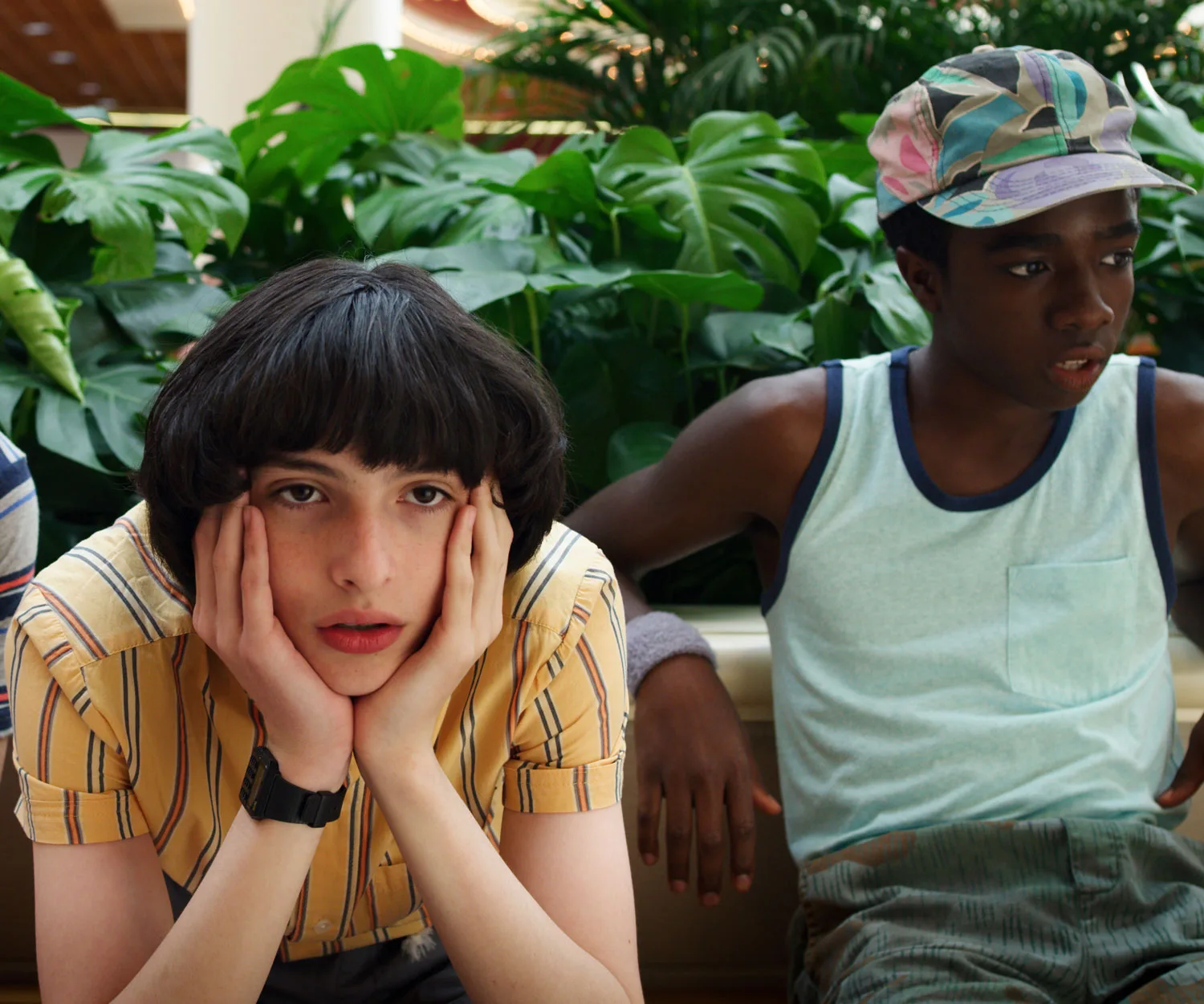 Two teenagers sitting in a mall; one looks bored, hands on face, the other gazes off, wearing a colorful cap.