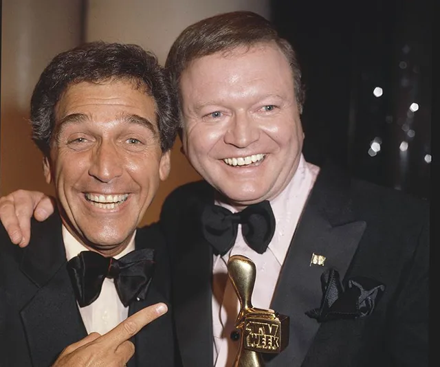 Two men in tuxedos smile and pose together, one holding a golden TV Week award.