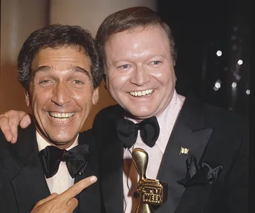 Two men in tuxedos smile and pose together, one holding a golden TV Week award.