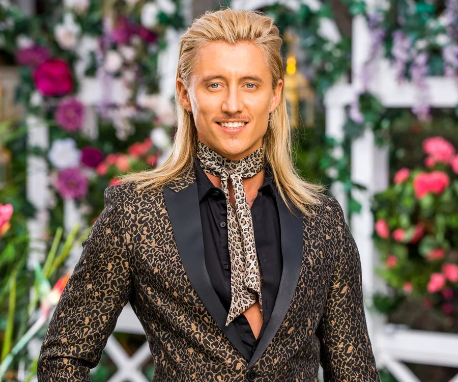 Man with long blond hair, leopard print suit, and scarf, standing in a garden with flowers in the background.