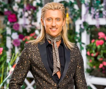 Man with long blond hair, leopard print suit, and scarf, standing in a garden with flowers in the background.