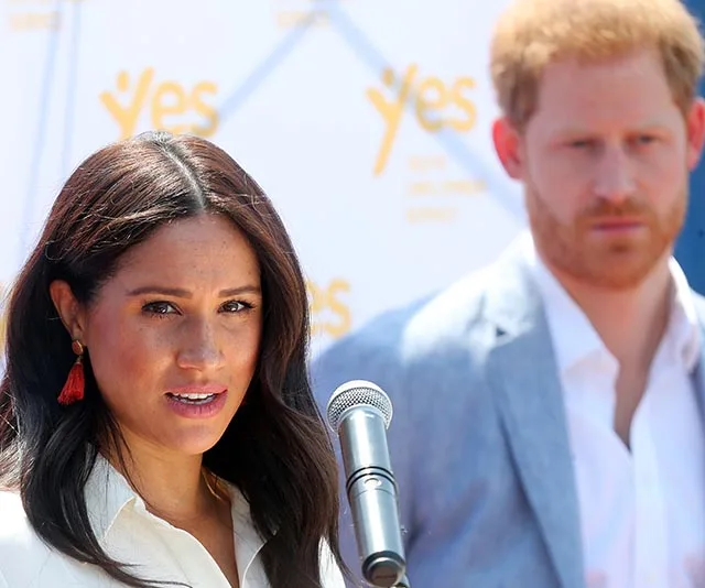 Meghan Markle speaking at a podium, Prince Harry standing behind with a focused expression.