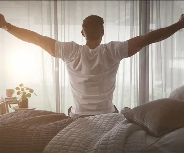 Man stretching arms while sitting on bed, morning light filters through curtains.