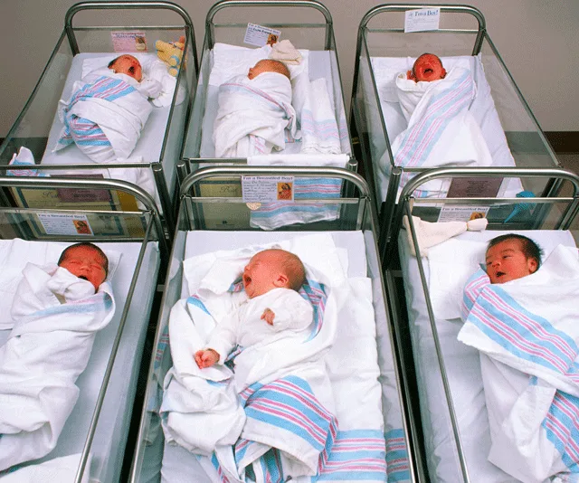 Hospital nursery with six newborn babies in bassinets, wrapped in striped blankets.
