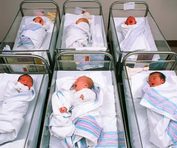 Hospital nursery with six newborn babies in bassinets, wrapped in striped blankets.