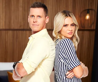 Two people stand back-to-back with arms crossed, looking serious, in a warmly lit room with wooden walls.