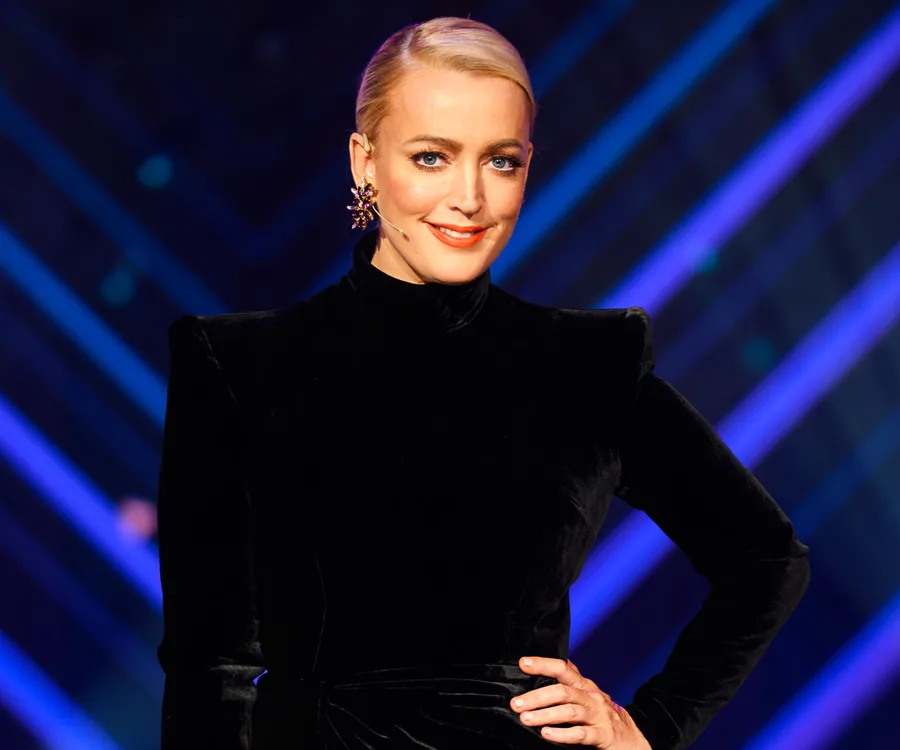 A person in a black outfit, smiling on a stage with a patterned blue and purple background.