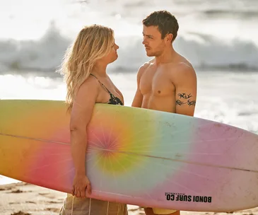 A man and woman stand on a beach with a rainbow-colored surfboard, facing each other under the sun.