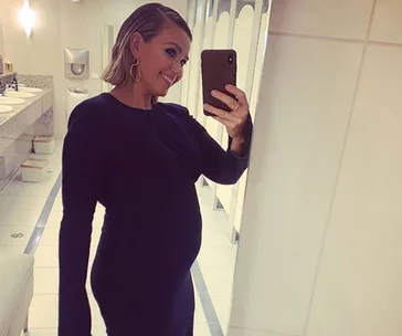 A woman in a black dress takes a mirror selfie in a bathroom, smiling.