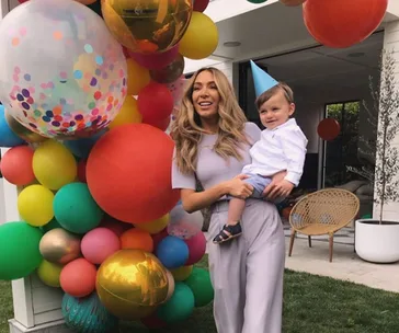 Woman holding a child in a blue party hat, surrounded by colorful balloons at an outdoor celebration.