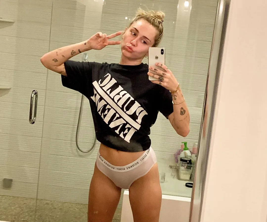 Person taking a mirror selfie in black T-shirt with text, making a peace sign and a playful expression in a bathroom.