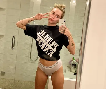 Person taking a mirror selfie in black T-shirt with text, making a peace sign and a playful expression in a bathroom.