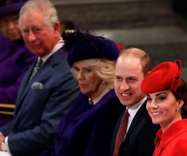 A group of elegantly dressed individuals seated in a row, smiling, and wearing formal attire and hats.