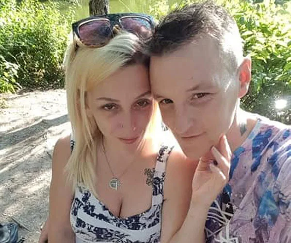 A couple poses closely together in a sunny outdoor setting, surrounded by greenery.
