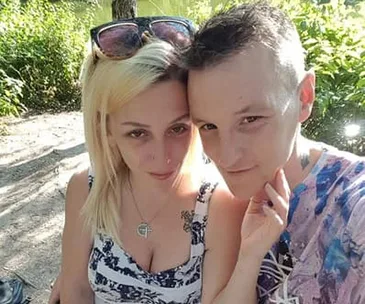 A couple poses closely together in a sunny outdoor setting, surrounded by greenery.