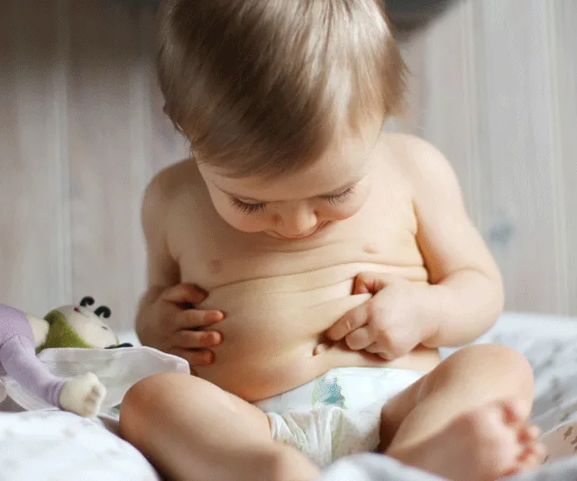 Baby in diaper looks at and touches belly button while sitting on a bed with a toy nearby.
