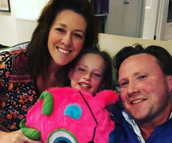 Three people smiling together, with a child holding a plush pink toy.