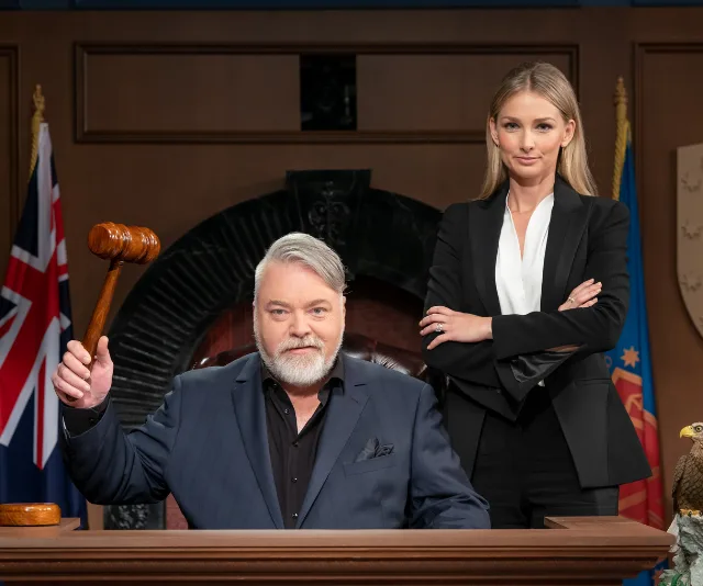 A judge holds a gavel while a woman stands beside him in a courtroom setting, with flags in the background.