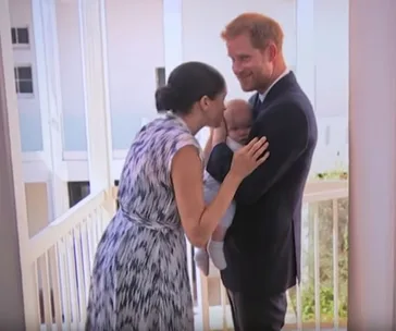 Parents holding a baby on a balcony, woman in patterned dress kisses baby held by man in a suit, smiling.