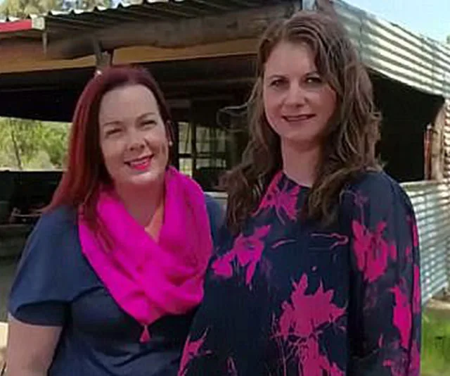 Two women smiling outdoors, one with a pink scarf, the other in a floral blouse, standing in front of a corrugated shed.